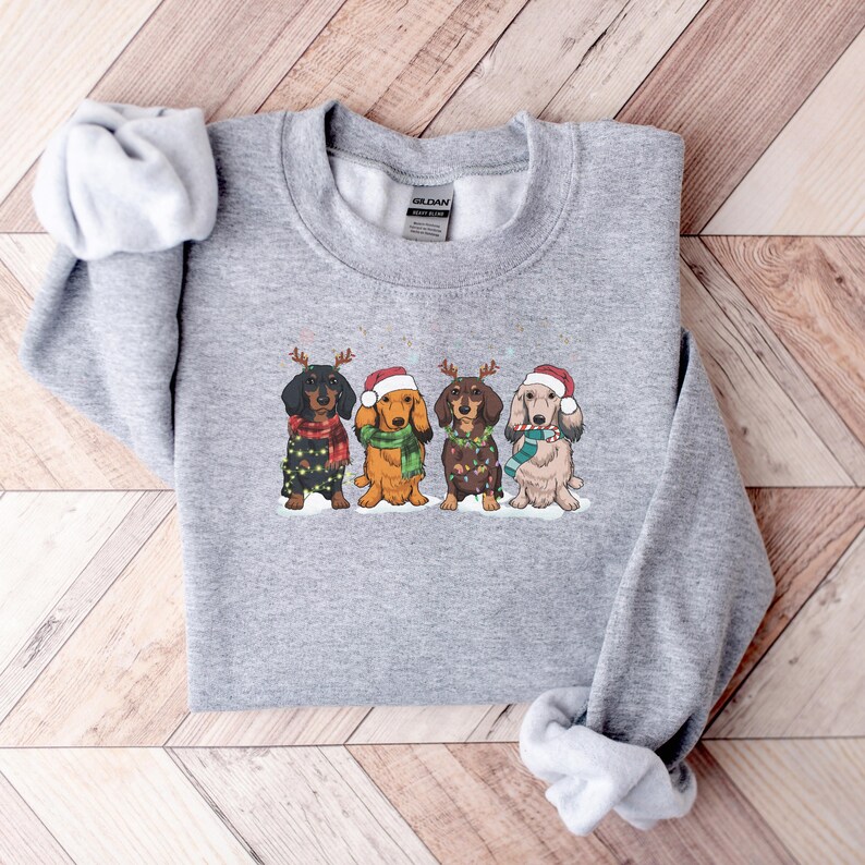 Op de afbeelding: Grijs sweatshirt met een kerstthema ontwerp met vier teckels die kerstmanmutsen en rendiergewei dragen. De honden dragen sjaals en hebben lichtjes om hun nek.