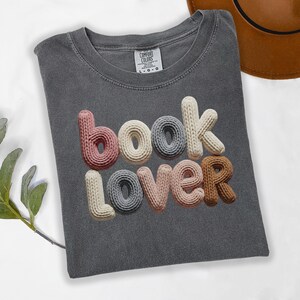 Könnte beinhalten: Ein graues T-Shirt mit dem Schriftzug "book lover" in einem 3D-Strickdesign. Die Buchstaben sind in Rosa-, Creme-, Grau- und Brauntönen gehalten. Das Shirt ist gefaltet, und ein brauner Hut ist teilweise im Hintergrund zu sehen.