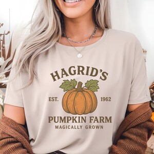 May include: Beige t-shirt with the text "Hagrid's Pumpkin Farm" and a pumpkin graphic. The design includes the text "Est. 1962" and "Magically Grown". The shirt is a neutral color and has a round neckline.