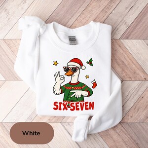May include: White sweatshirt featuring a cartoon goose wearing sunglasses and a Santa hat. The goose is giving an 'okay' hand gesture and wearing a green Christmas sweater. The text "SIX-SEVEN" is printed in red below the goose.