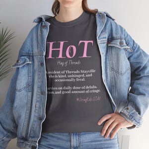 Op de afbeelding: Een donkergrijs t-shirt met de roze tekst HoT en de witte tekst Hag of Threads, A resident of Threads Stayville who is kind, unhinged, and occasionally feral. Survives on daily dose of delulu, casm, and good amount of cringe. #StrayKidsSTAY.