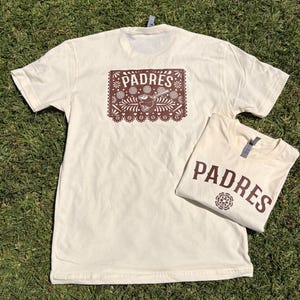 May include: Two cream-colored t-shirts with brown text and graphics. One shirt has a large design on the back with the word "PADRES" and a floral design. The other shirt is folded and has "PADRES" printed on the front.