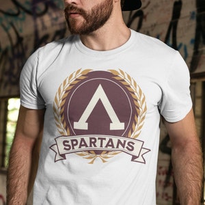 Könnte beinhalten: Ein weißes T-Shirt mit einem braunen und goldenen Spartaner-Logo. Das Logo zeigt einen Schild mit einem stilisierten Buchstaben A und einem Lorbeerkranz. Der Text "SPARTANS" steht auf einem Banner unter dem Schild.