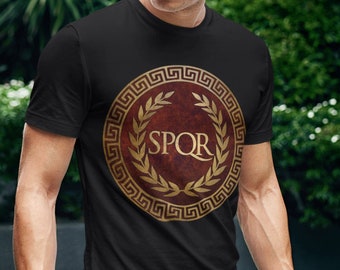 Puede incluir: Camiseta negra con un gráfico dorado y rojo de una corona de laurel con el texto "SPQR" en su interior.