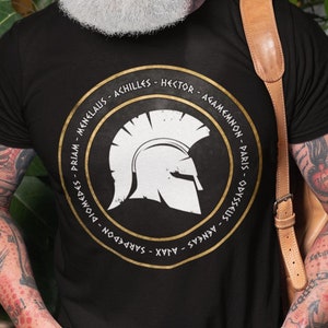 Op de afbeelding: Een zwart T-shirt met een gouden cirkelvormige afbeelding met een witte Spartaanse helm en de namen van beroemde Griekse krijgers in gouden letters. De namen zijn: Menelaos, Achilles, Hector, Agamemnon, Paris, Odysseus, Diomedes, Priamus, Sarpedon, Ajax, Aeneas.