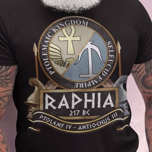 May include: A black t-shirt with a graphic design of a circular emblem featuring a winged ankh and a trident. The emblem is divided in half, with the top half representing the Ptolemaic Kingdom and the bottom half representing the Seleucid Empire. The text "RAPHIA 217 BC PTOLEMY IV - ANTIOCHUS III" is printed below the emblem.