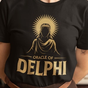 May include: A black t-shirt with a gold graphic of a woman with a halo and the text "Oracle of Delphi".
