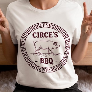 May include: White t-shirt featuring a circular design with the text "CIRCE'S BBQ" and a pig illustration. The design is in a dark maroon color. The shirt is made of a soft material.