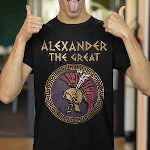 Peut inclure: T-shirt noir avec un texte doré qui dit "ALEXANDER THE GREAT" et un graphique d'un casque doré et rouge avec une plume à l'intérieur d'un cercle doré avec un motif de clé grecque.