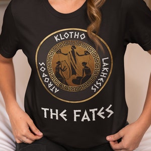 May include: Black t-shirt with a gold circle graphic featuring three figures and the text "KLOTHO, LAKHESIS, ATROPOS" in Greek. The text "THE FATES" is printed below the circle.