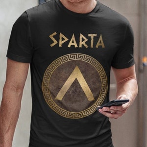 Peut inclure: Un t-shirt noir avec un graphique doré d'un bouclier spartiate avec le mot "SPARTA" au-dessus.