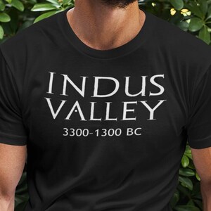 May include: Black t-shirt with the text "INDUS VALLEY" and the dates "3300-1300 BC" in white. The shirt is short-sleeved and appears to be made of cotton. The person wearing the shirt is outdoors, in front of green foliage.
