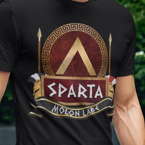 May include: Black t-shirt featuring a gold and red design with the word "SPARTA" and the phrase "MOLON LABE". The shirt has a circular emblem with a gold lambda symbol and spear details. The shirt is worn with blue jeans.
