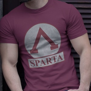 Puede incluir: Una camiseta granate con un círculo blanco y un triángulo rojo en su interior. El texto "SPARTA" está debajo del círculo.