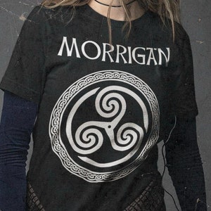 May include: A woman wearing a black t-shirt with a white Celtic knot design and the word "MORRIGAN" printed on it. She is also wearing a plaid skirt and black leggings.