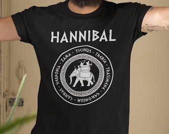Puede incluir: Un hombre con un gorro negro y una camiseta negra con un gráfico circular blanco que dice "HANNIBAL" en mayúsculas. El gráfico presenta un elefante blanco con jinetes y texto en un patrón circular alrededor del elefante.