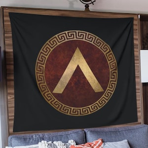 Puede incluir: Un tapiz negro con un escudo dorado y rojo que presenta una punta de flecha estilizada. El escudo está rodeado por un patrón de llave griega dorada. El tapiz está colgado en una pared sobre un sofá gris con almohadas.