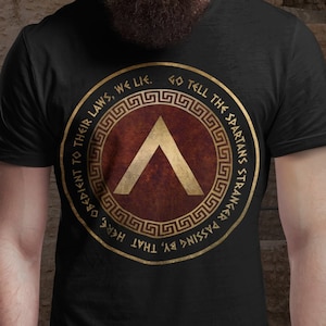 Puede incluir: Camiseta negra con un gráfico de escudo espartano dorado. El escudo presenta un chevron rojo y el texto "GO TELL THE SPARTANS THAT PASSING BY MISSED THAT WE LIE. OBEIDIENT TO THEIR LAWS, WE LIE."
