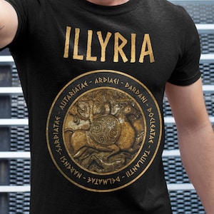 Puede incluir: Una camiseta negra con un gráfico dorado de un diseño circular que presenta una imagen estilizada de un guerrero con un escudo y la palabra "ILLYRIA" en letras doradas.