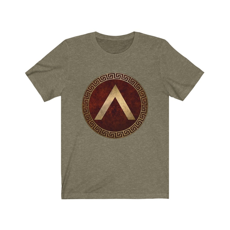 Puede incluir: Una camiseta gris jaspeada con un dise&ntilde;o de escudo griego rojo y dorado. El escudo presenta un gal&oacute;n dorado dentro de un c&iacute;rculo dorado con un borde de clave griega.
