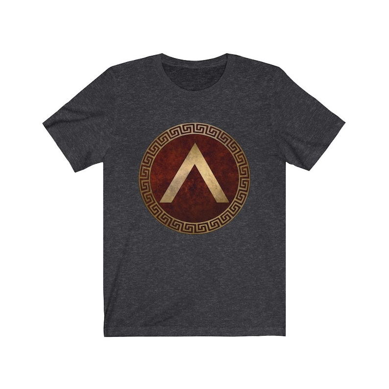 Puede incluir: Una camiseta gris oscuro jaspeada con un dise&ntilde;o de escudo de guerrero griego en oro y rojo. El escudo presenta un s&iacute;mbolo de chevron dorado dentro de un c&iacute;rculo dorado con fondo rojo. El c&iacute;rculo est&aacute; rodeado por un patr&oacute;n de llave griega dorada.