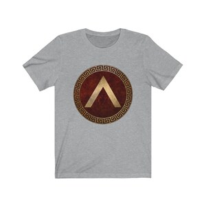 Puede incluir: Una camiseta gris con un escudo rojo y dorado que presenta una punta de flecha dorada estilizada. El escudo est&aacute; rodeado por un patr&oacute;n de llave griega dorada.