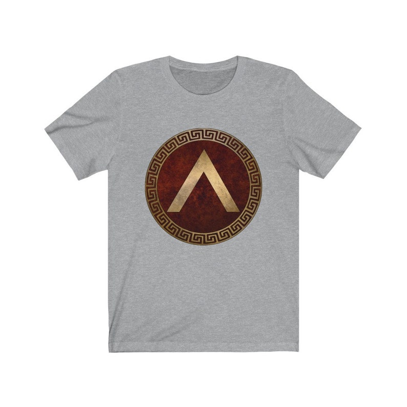 Puede incluir: Una camiseta gris con un escudo rojo y dorado que presenta una punta de flecha dorada estilizada. El escudo est&aacute; rodeado por un patr&oacute;n de llave griega dorada.