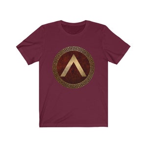 Puede incluir: Una camiseta granate con un dise&ntilde;o de llave griega dorada alrededor de un escudo rojo con un gal&oacute;n dorado en el interior.