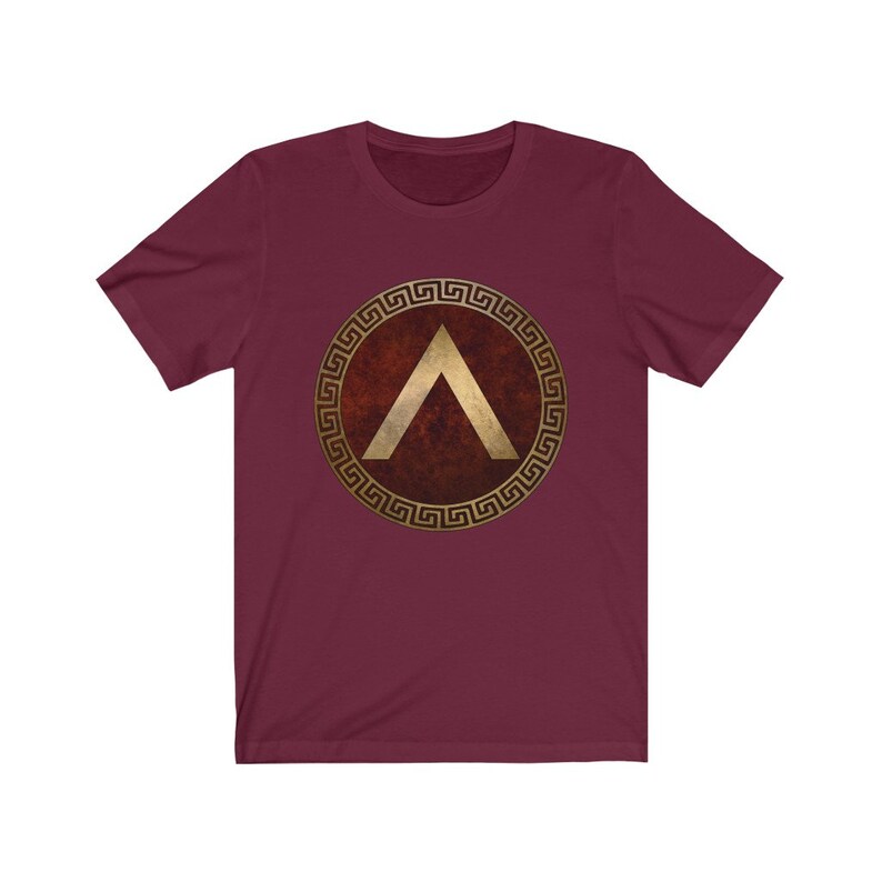 Puede incluir: Una camiseta granate con un dise&ntilde;o de llave griega dorada alrededor de un escudo rojo con un gal&oacute;n dorado en el interior.