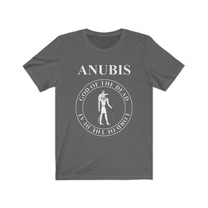 Op de afbeelding: Een donkergrijs T-shirt met een witte afbeelding van Anubis, de Egyptische god van de dood. De afbeelding toont een cirkel met de tekst "God van de Doden" en "Heer van de Duat" rond de afbeelding van Anubis.