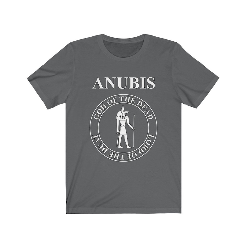 Op de afbeelding: Een donkergrijs T-shirt met een witte afbeelding van Anubis, de Egyptische god van de dood. De afbeelding toont een cirkel met de tekst "God van de Doden" en "Heer van de Duat" rond de afbeelding van Anubis.