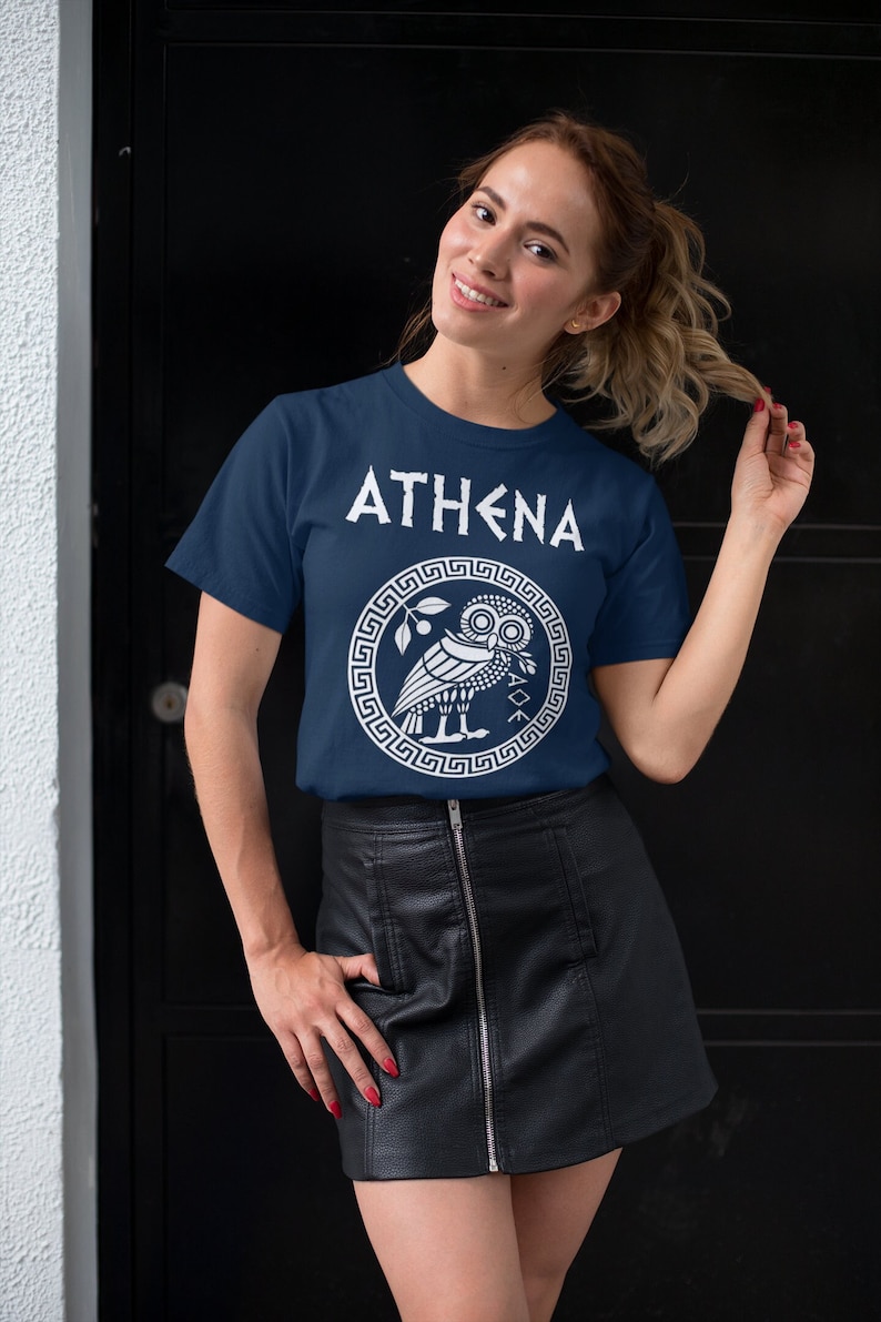 May include: A navy blue t-shirt with a white graphic of an owl and the text "ATHENA" in a stylized font. The owl is surrounded by a white Greek key border.