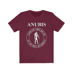 Op de afbeelding: Een maroon t-shirt met een witte afbeelding van Anubis, de Egyptische god van de dood. De afbeelding bevat de tekst "Anubis, God van de Doden, Heer van de Duat".