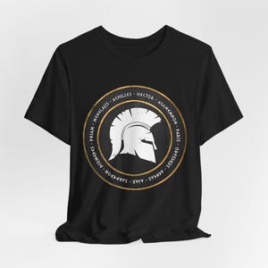 Op de afbeelding: Zwart T-shirt met een gouden cirkelvormige afbeelding met een witte Spartaanse helm en de namen van beroemde Griekse krijgers in gouden letters.