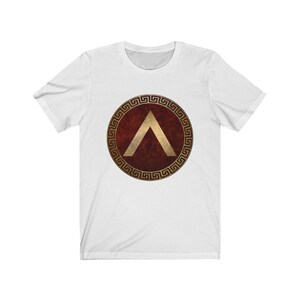 Puede incluir: Camiseta blanca con un dise&ntilde;o de escudo griego rojo y dorado. El escudo presenta un gal&oacute;n dorado dentro de un c&iacute;rculo dorado con un borde de clave griega.