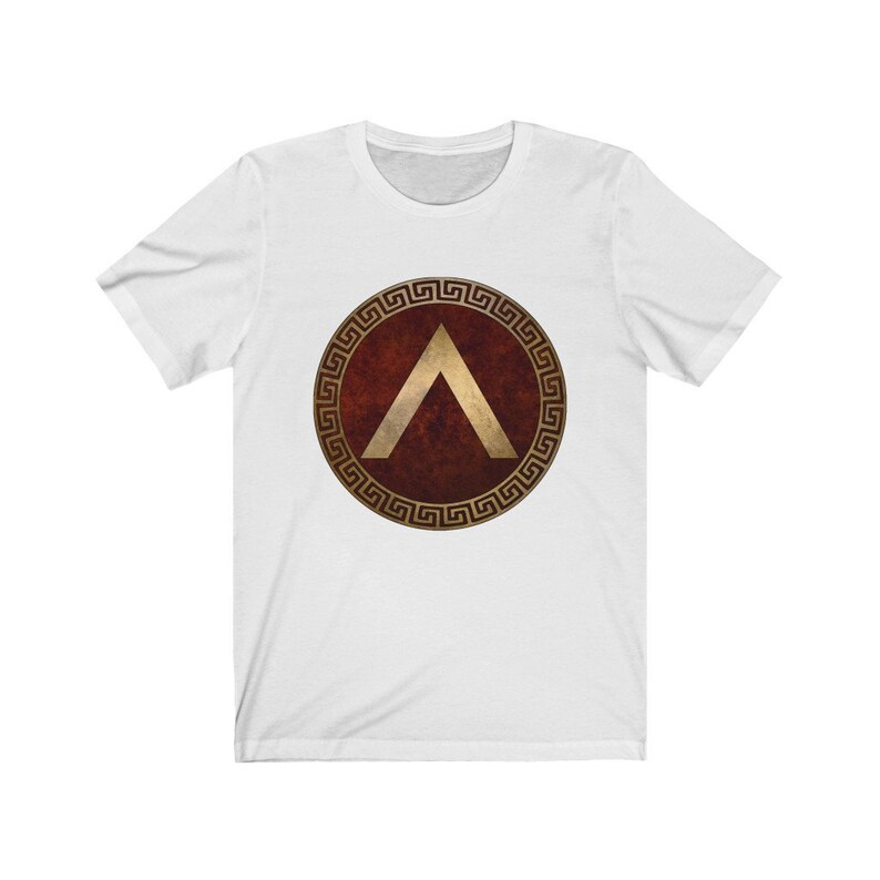 Puede incluir: Camiseta blanca con un dise&ntilde;o de escudo griego rojo y dorado. El escudo presenta un gal&oacute;n dorado dentro de un c&iacute;rculo dorado con un borde de clave griega.
