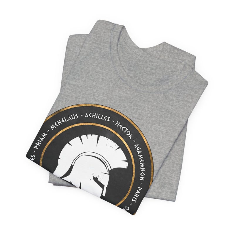 Op de afbeelding: Grijs T-shirt met een zwarte en gouden cirkelvormige afbeelding met een witte Spartaanse helm en de namen van beroemde Trojaanse oorlogspersonages in Grieks schrift: Priamus, Menelaos, Achilles, Hector, Agamemnon, Paris.