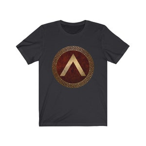 Puede incluir: Una camiseta gris oscuro con un dise&ntilde;o circular dorado y rojo. El dise&ntilde;o presenta un borde dorado en forma de llave griega y una forma de chevron dorada sobre un fondo rojo.