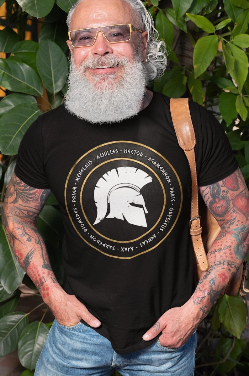 Op de afbeelding: Een zwart T-shirt met een gouden cirkelvormige afbeelding met een witte Spartaanse helm en de namen van beroemde Griekse krijgers in gouden letters. De namen zijn: Menelaos, Achilles, Hector, Agamemnon, Paris, Odysseus, Diomedes, Priamus, Sarpedon, Ajax, Aeneas.