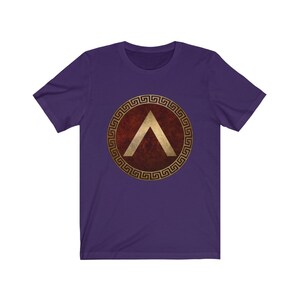 Puede incluir: Camiseta de manga corta de color morado con un dise&ntilde;o de escudo dorado y rojo. El escudo presenta un gal&oacute;n dorado dentro de un c&iacute;rculo dorado con un borde de clave griega.
