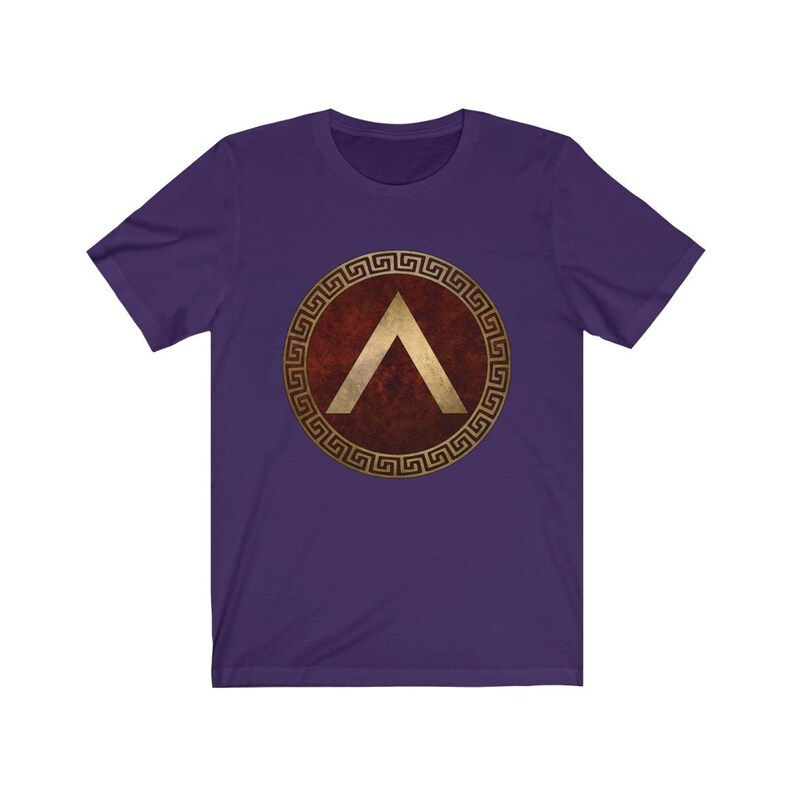 Puede incluir: Camiseta de manga corta de color morado con un dise&ntilde;o de escudo dorado y rojo. El escudo presenta un gal&oacute;n dorado dentro de un c&iacute;rculo dorado con un borde de clave griega.
