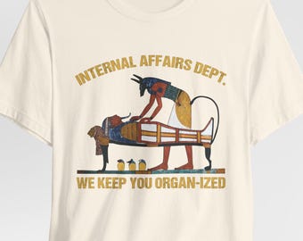 May include: Cream-colored t-shirt featuring a graphic of an Egyptian scene with the text "INTERNAL AFFAIRS DEPT." above and "WE KEEP YOU ORGAN-IZED" below. The graphic depicts a figure attending to a sarcophagus.