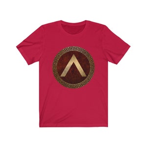 Puede incluir: Camiseta roja con un dise&ntilde;o de escudo griego dorado que presenta una punta de flecha estilizada.