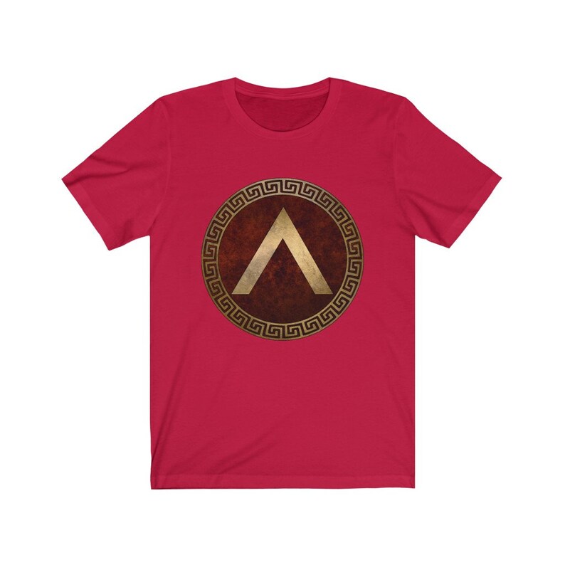 Puede incluir: Camiseta roja con un dise&ntilde;o de escudo griego dorado que presenta una punta de flecha estilizada.