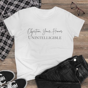 以下が含まれることがあります： 白いTシャツに黒い文字で「異議あり、ご清聴ありがとうございます。理解不能」と書かれています。Tシャツは木製の表面に、チェックシャツ、ジーンズ、スニーカーと一緒に置かれています。
