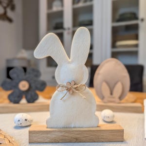 Könnte beinhalten: Osterdekoration mit einem weißen Filzhasen auf einem Holzsockel, verziert mit einer Schnur-Schleife und einer kleinen Holzblume. Enthält eine graue Blume und ein gesprenkeltes Ei. Der Hase ist etwa 15 cm hoch.