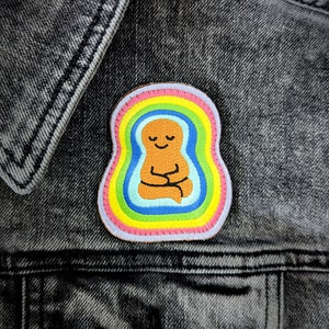 May include: A colorful embroidered patch featuring a brown figure meditating inside a rainbow. The figure has a smiling face and is sitting in a lotus position.