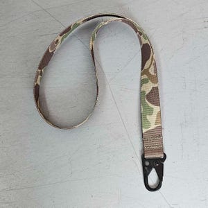 May include: A fabric lanyard featuring a camouflage pattern in shades of green, brown, and tan. The strap is constructed from woven material and includes a black metal lobster claw clasp attached at the bottom end with reinforced stitching.