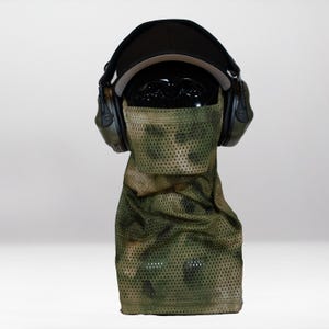Op de afbeelding: Een zwarte honkbalpet, donkergroene gehoorbescherming en een camouflage-achtig mesh gezichtsmasker dat het onderste deel van het gezicht bedekt. Het masker heeft een geperforeerde textuur in olijfgroen, bruin en beige op een paspophoofd.