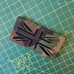 May include: A rectangular patch featuring a camouflage pattern in shades of green, brown, and black. The patch displays a laser-cut Union Jack design, revealing a solid black layer underneath. The edges are stitched with matching thread.